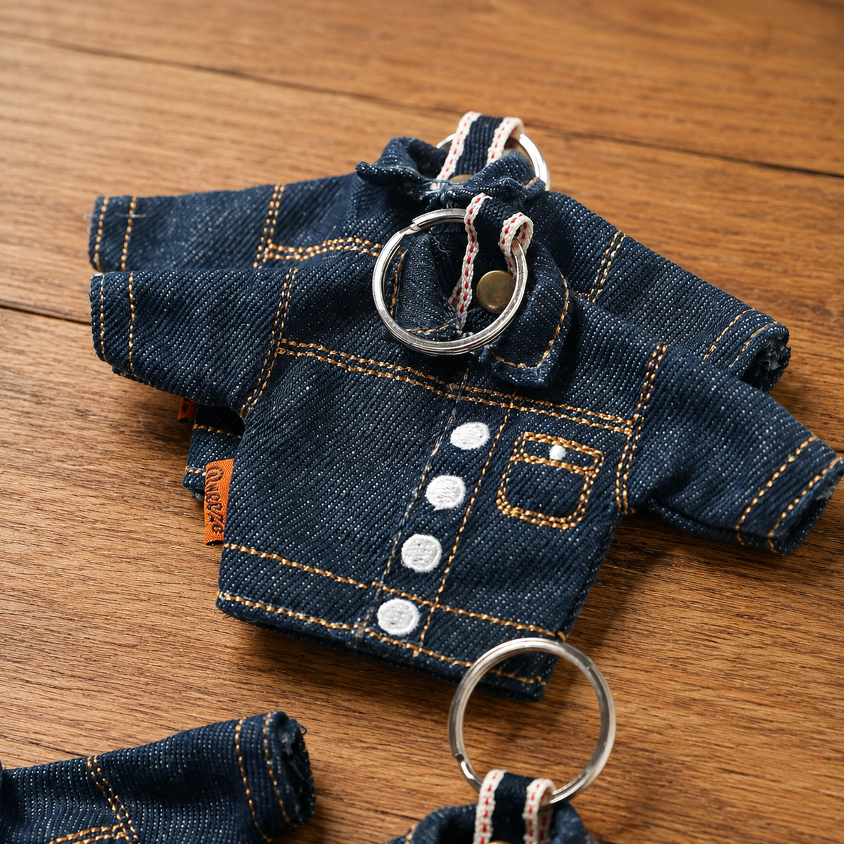A miniature denim-style keychain outfit for small pets, featuring a dark blue denim shirt with gold contrast stitching, white buttons, and silver metal key rings, displayed on a wooden surface.