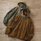 Two hooded pullover jackets laid flat on a light wood floor. The front jacket is a warm camel-brown with a half-zip neckline, drawstrings, and zip pockets.