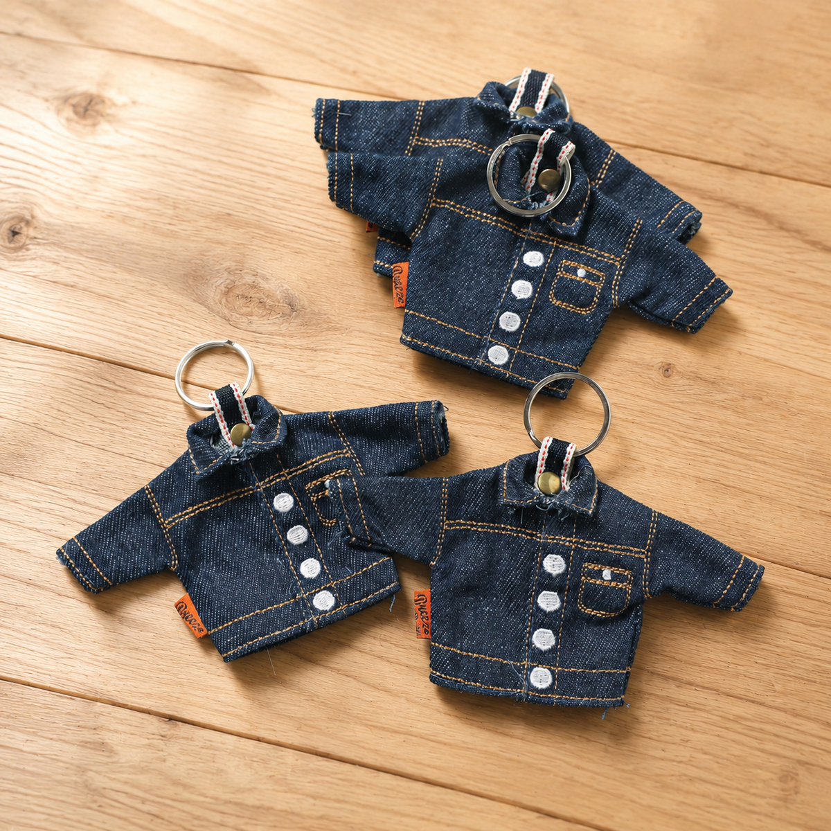 Three miniature denim jacket keychains with silver metal rings, displayed on a light brown wooden surface.